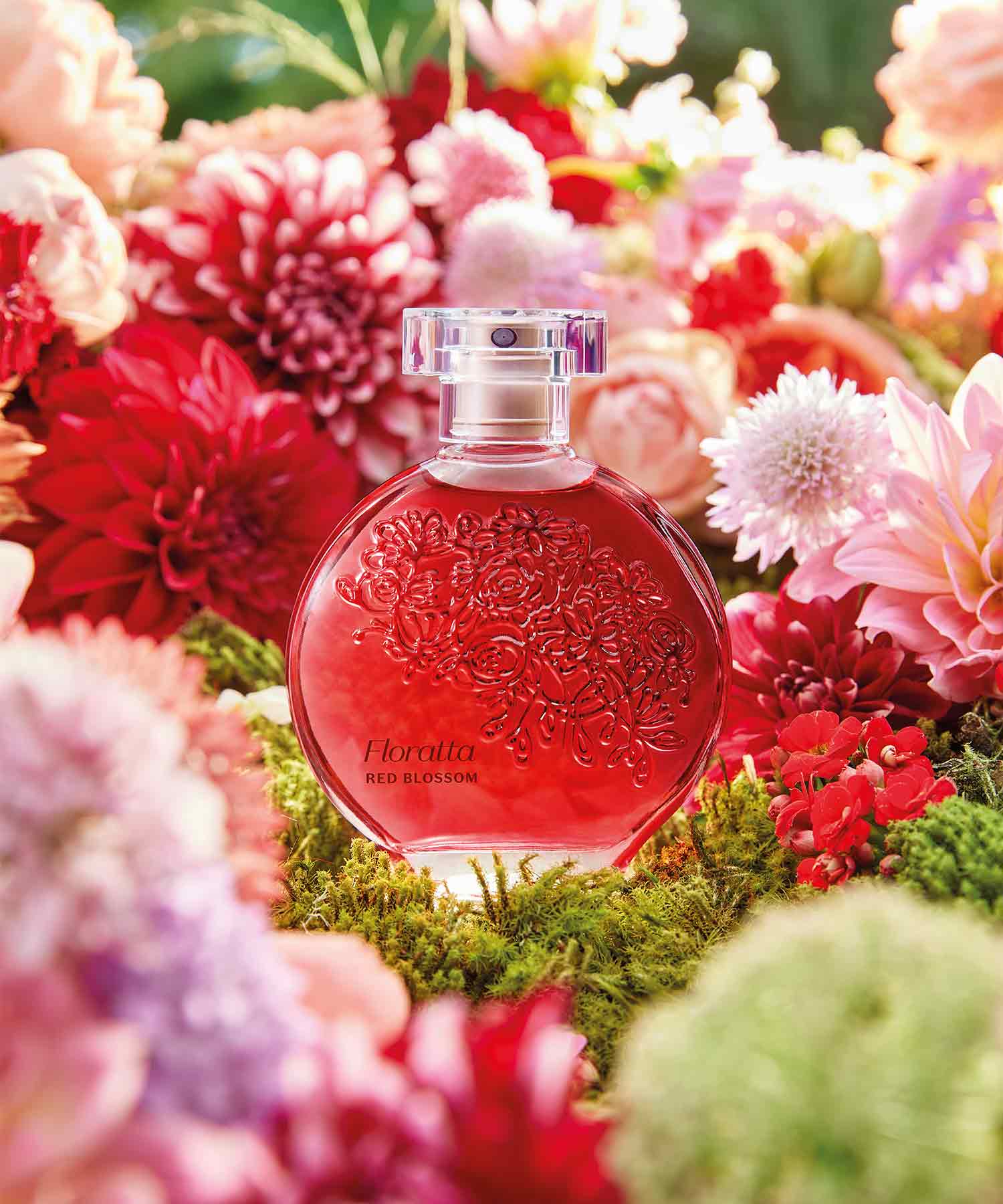 Frasco de perfume Floratta Red num ambiente floral alusivo ao Dia da Mãe. A embalagem é em vidro transparente, com um líquido vermelho intenso dentro e uma tampa transparente. O rótulo tem o nome do perfume em letras elegantes e o desenho floral no frasco.