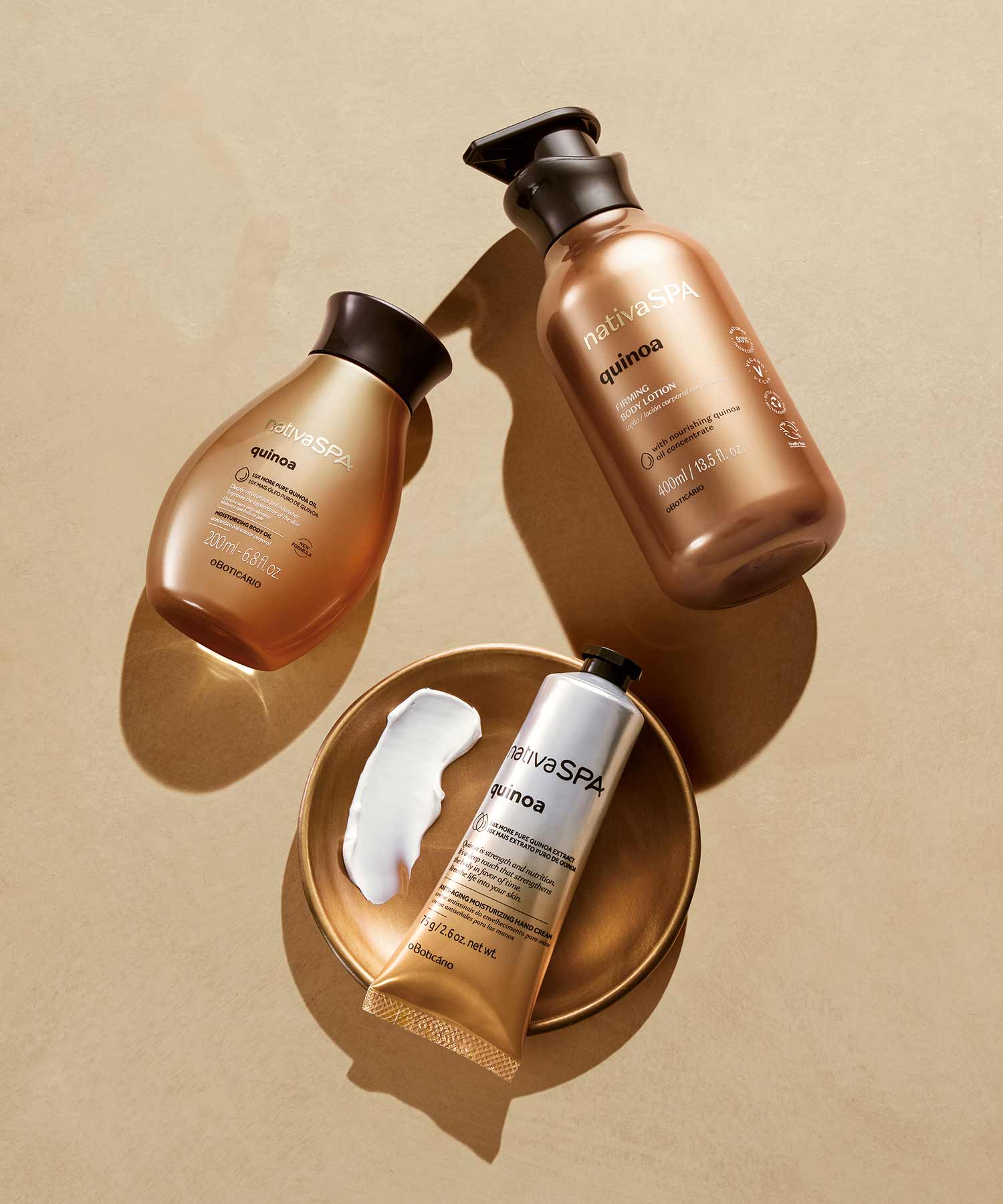 Três produtos da marca Nativa SPA, da linha Quinoa. Um creme hidratante corporal, um óleo de banho e um creme de mãos de cor acastanha e dourada. Os três produtos estão deitados num fundo claro, dando um design elegante.